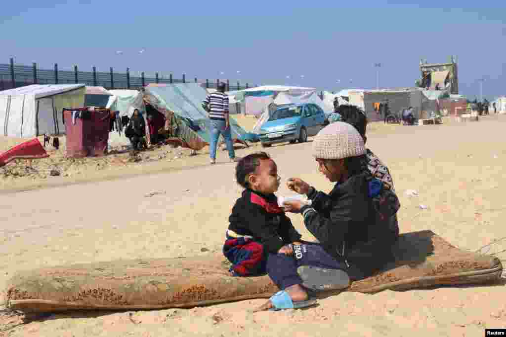 Displaced Palestinian girl, who fled her house due to Israeli strikes, feeds her brother at a tent camp, near the border with Egypt, in Rafah in the southern Gaza Strip.
